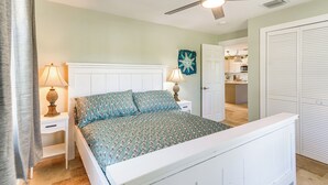 Ein Schlafzimmer mit einem weißen Bett, zwei Nachttischen mit Lampen, einem Deckenventilator und Blick in die Küche.