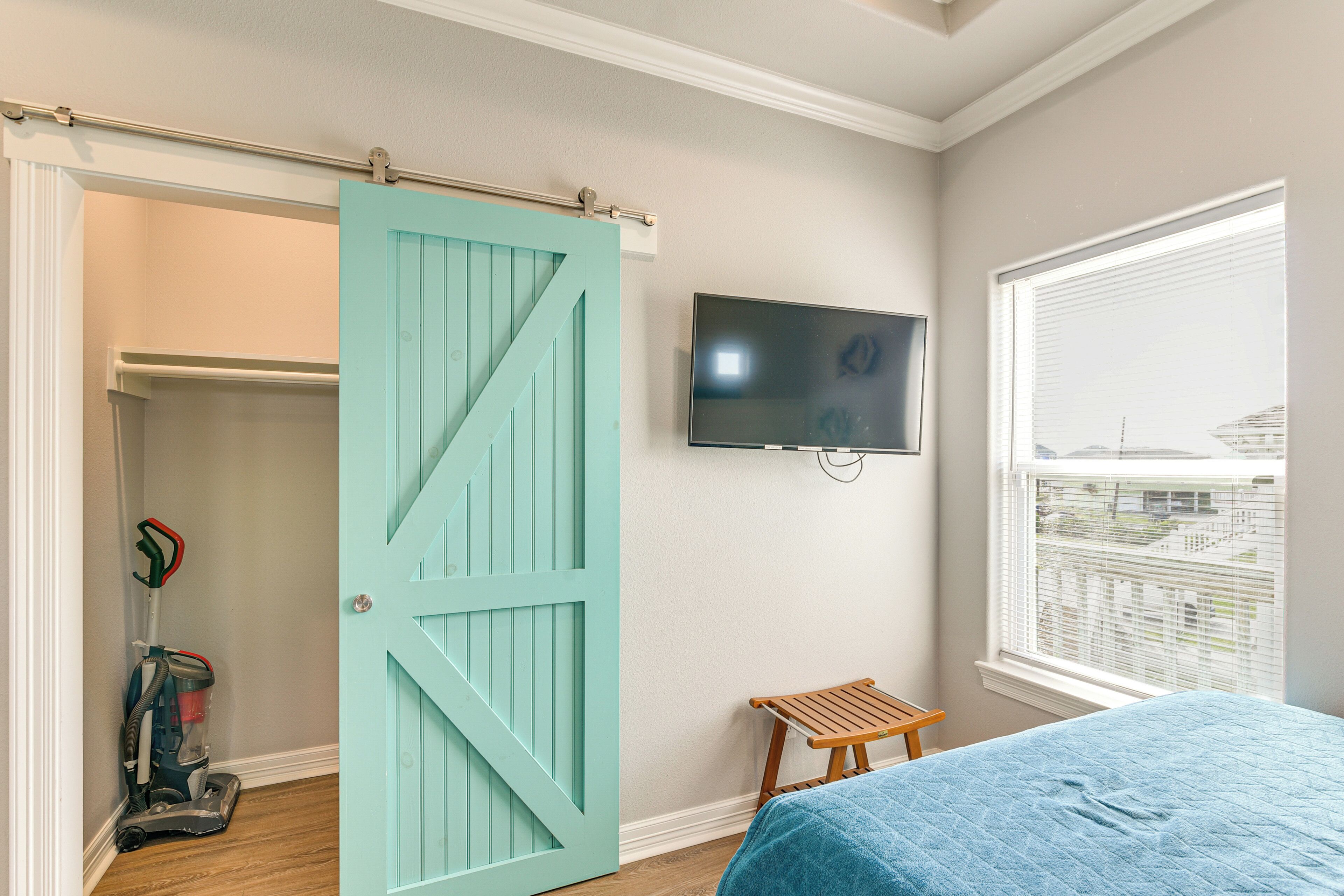 Une chambre avec une porte coulissante de couleur turquoise, un lit recouvert d’une couverture bleue, un téléviseur fixé au mur et une fenêtre avec des stores.