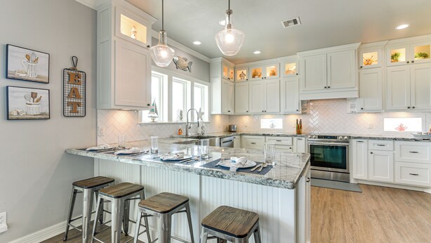 Una cocina moderna con armarios blancos, una isla central con taburetes, iluminación colgante y un protector contra salpicaduras de azulejos.