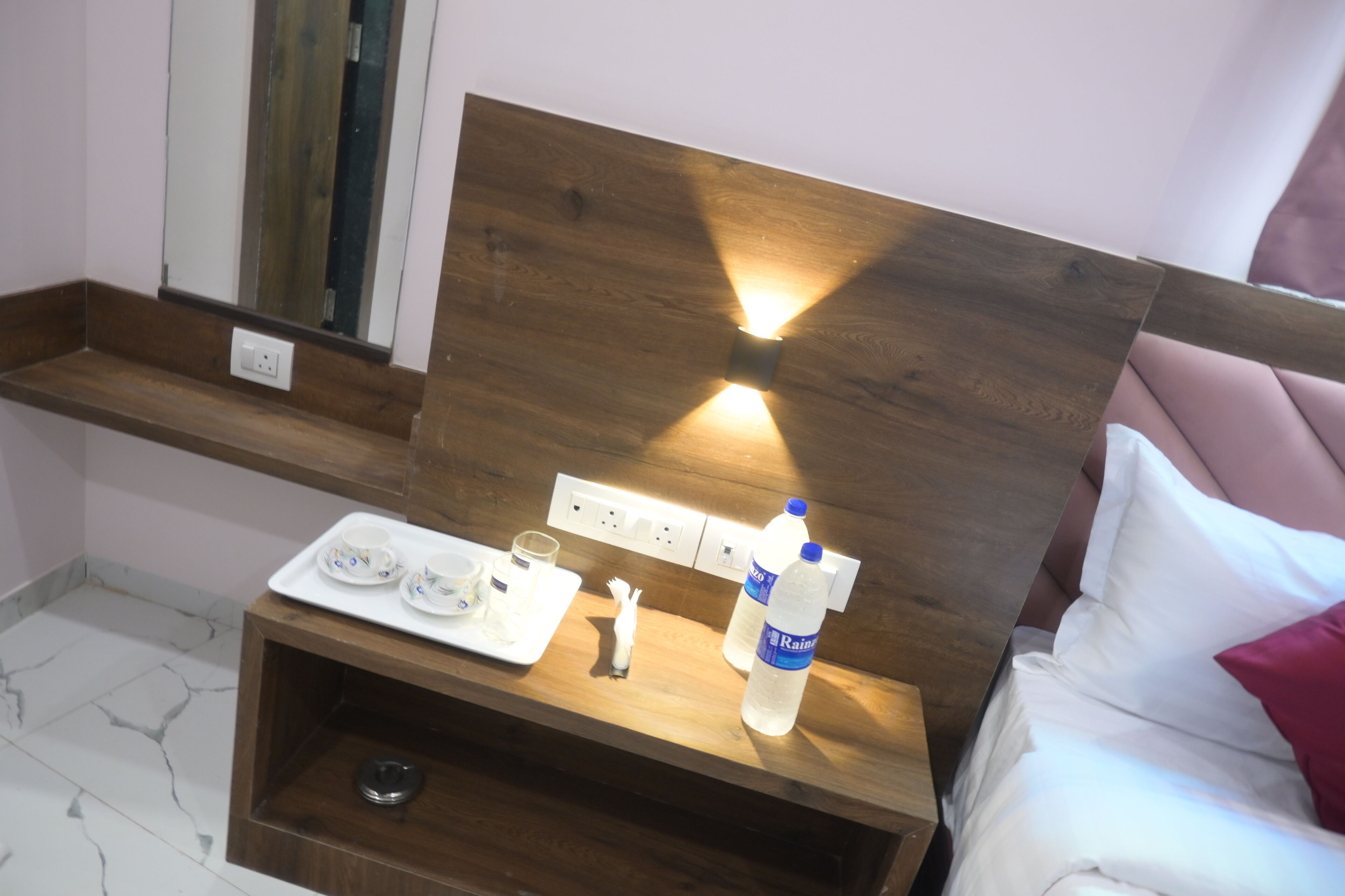 Une chambre d’hôtel avec une tête de lit en bois, une table de chevet sur laquelle se trouve un plateau avec du thé et des bouteilles d’eau, et un miroir au-dessus de la table.