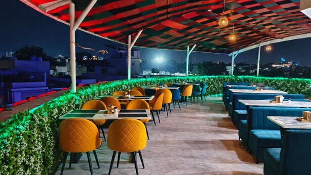An outdoor dining area with tables and chairs, green foliage, and a cityscape view in the background. - Hotel The Muffet (Indore)