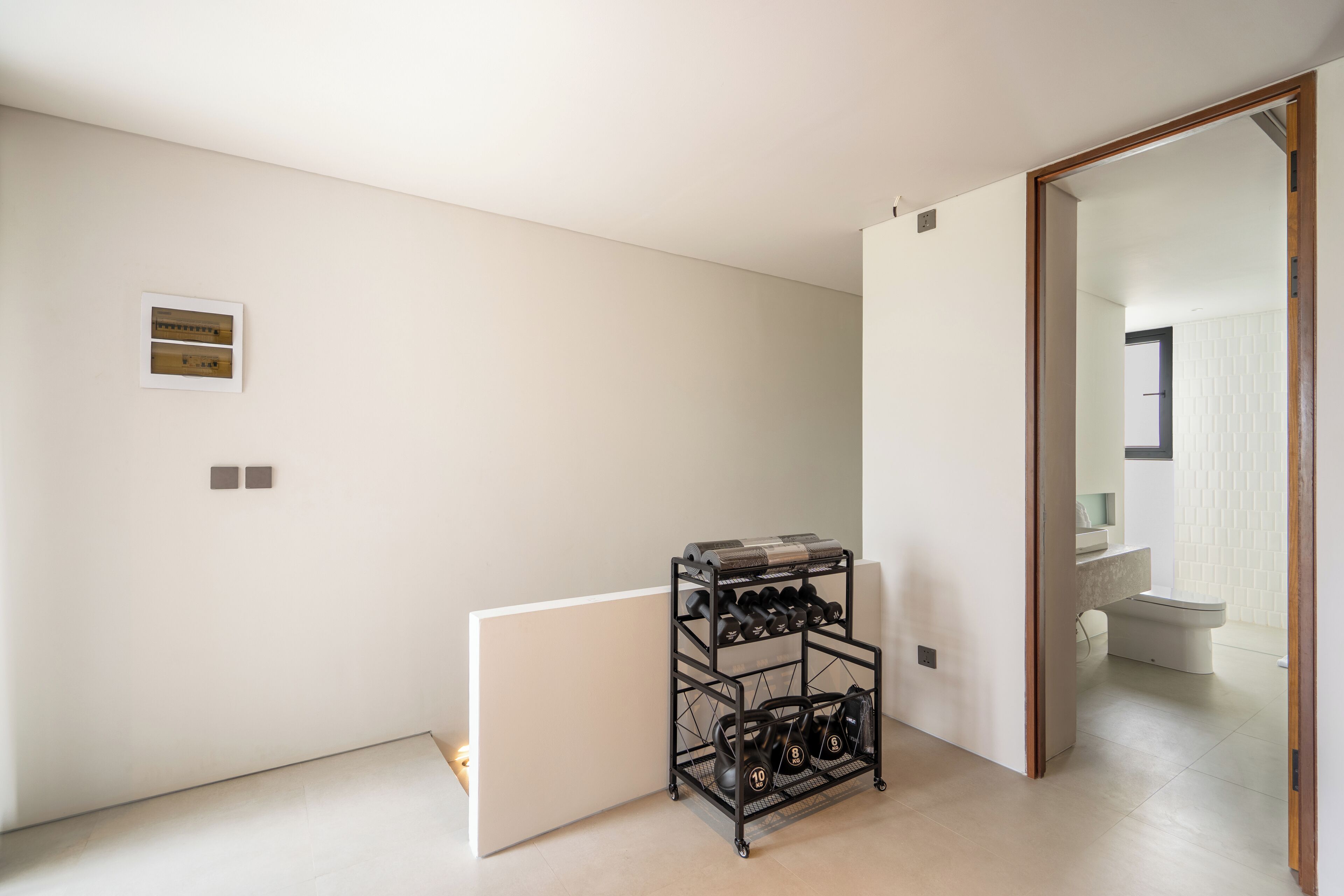 Habitación minimalista con una estantería de metal negro que contiene botellas de vino y mancuernas, un radiador blanco y una puerta que da a un baño con pared de azulejos blancos.