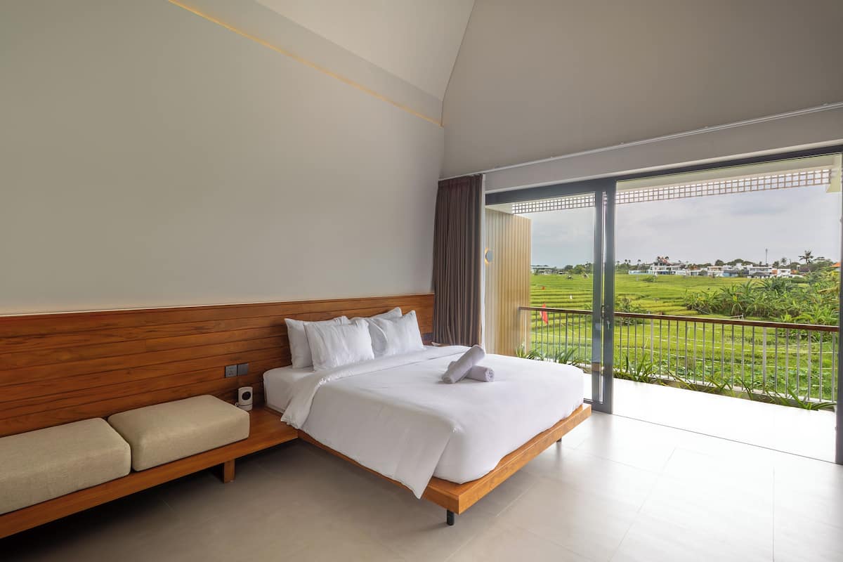 A modern hotel room with a large bed, a wooden bench, and a balcony with a view of a green landscape.