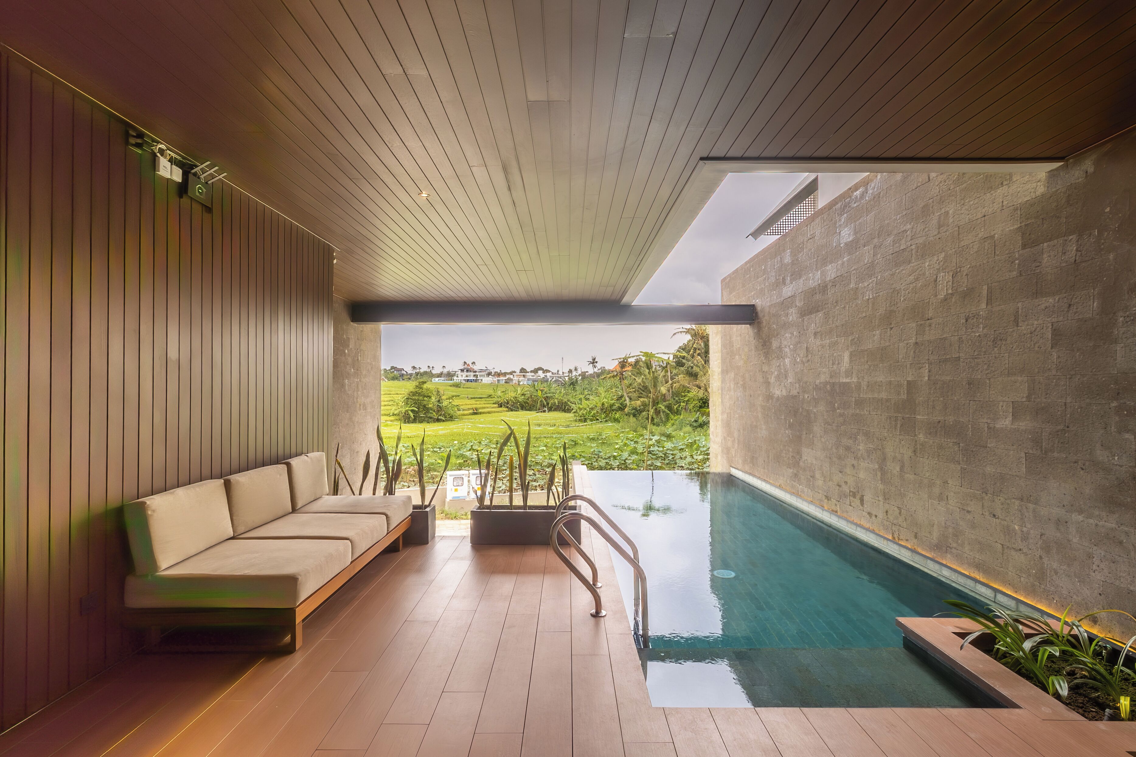 A modern indoor pool area with a wooden ceiling, a sofa, and a view of a lush landscape.