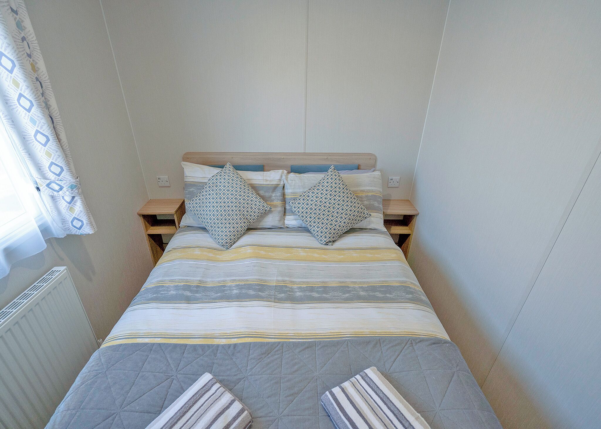 A small, neatly arranged bedroom with a bed, two pillows, a bedside table, and a radiator.