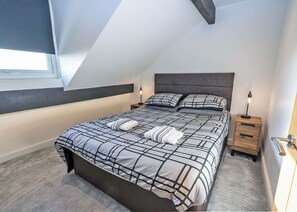 A bedroom with a sloped ceiling, a bed with a geometric-patterned bedspread, two bedside lamps, and a wooden nightstand. - 2 bedroom accommodation in Millom (Millom)