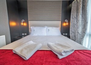 Uma cama de casal com roupa de cama branca, duas almofadas, um cobertor vermelho e duas toalhas.