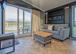 A modern living room with a sofa, a wooden coffee table, and a view of a deck and landscape through large glass doors. - Kestrel Retreat S-Pod 4 (Millom)