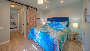 A bedroom with a bed, a ceiling fan, a nightstand, and a door leading to another room. - 2 Cozy Cottages | Private Yards, Fire Pit, Walkable Location in Spanish Village (Port Aransas)