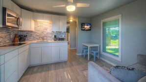 A modern kitchen with white cabinets, a microwave, and a built-in oven. There is a dining area with a small table and chairs, and a window with blinds. - 2 Cozy Cottages | Private Yards, Fire Pit, Walkable Location in Spanish Village (Port Aransas)