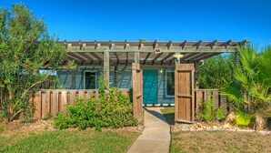 A wooden pergola with a pathway leading to a building with a green door. - 2 Cozy Cottages | Private Yards, Fire Pit, Walkable Location in Spanish Village (Port Aransas)