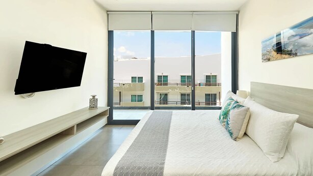 A modern hotel room with a large bed, a flat-screen TV, a ceiling fan, and a view of buildings through large windows. - 2BR condo w/ pools, gym & balcony, steps to beach (Playa del Carmen)