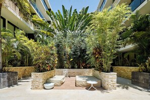 A courtyard with a seating area, surrounded by lush tropical plants and a modern building facade. - 2BR condo w/ pools, gym & balcony, steps to beach (Playa del Carmen)