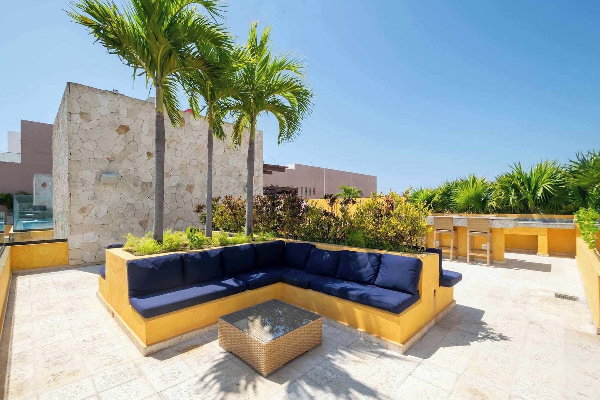 A modern outdoor seating area with a sectional sofa and a coffee table, surrounded by palm trees and plants.