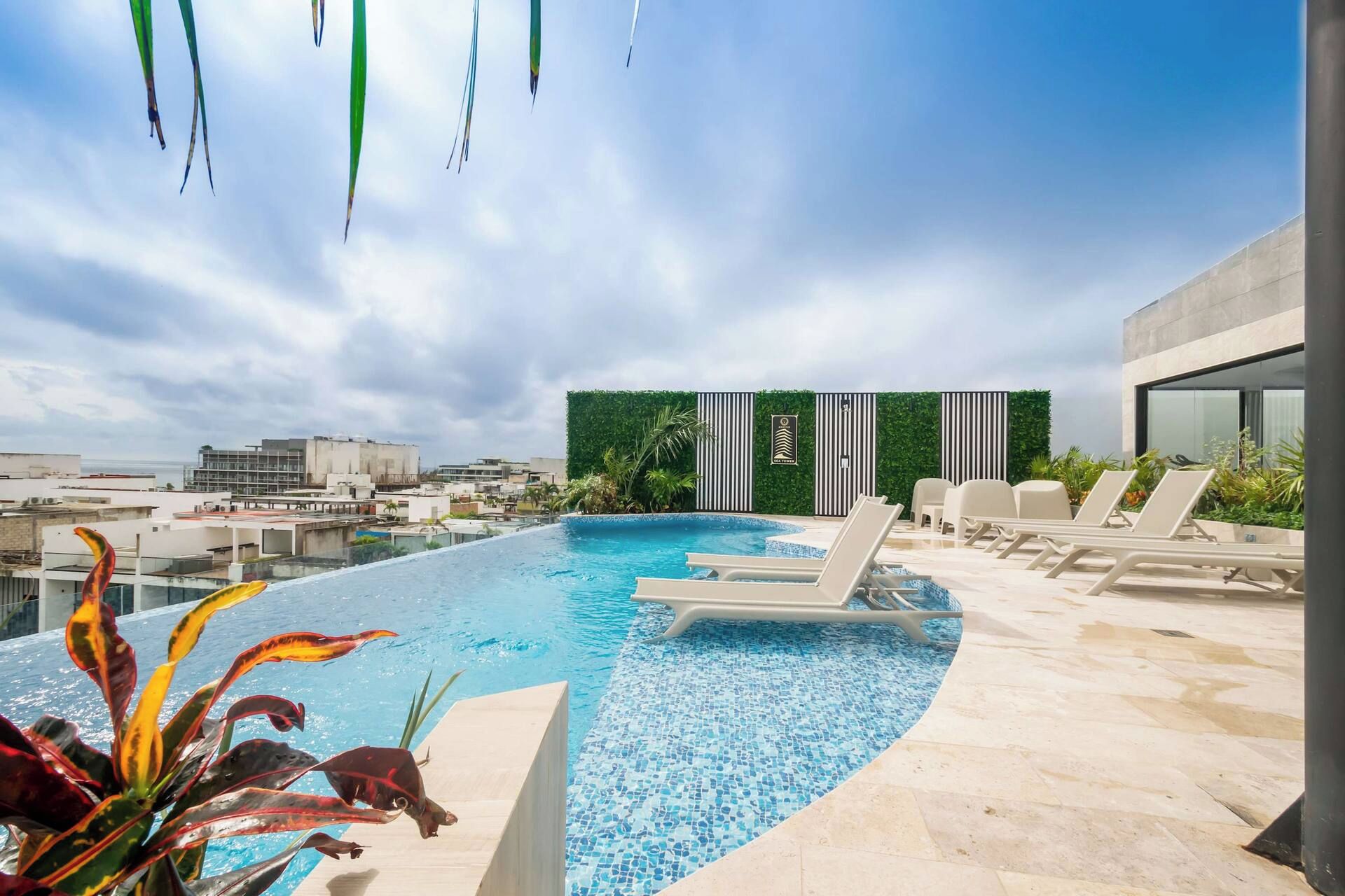 Ein Dachpool mit Infinity-Kante, Liegestühlen, einer grünen Wand mit vertikalen Streifen und einer Stadtkulisse im Hintergrund.