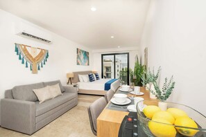 A modern hotel room with a sofa, a bed with a blue accent, a dining table with a bowl of lemons, and a large window. - Rooftop pool studio | steps to Coco Beach (Playa del Carmen)