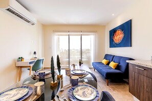 A modern living room with a glass dining table set for four, a blue sofa with yellow cushions, and a city view through large windows. - Veranto condo near 5th Ave w/ pool access (Playa del Carmen)