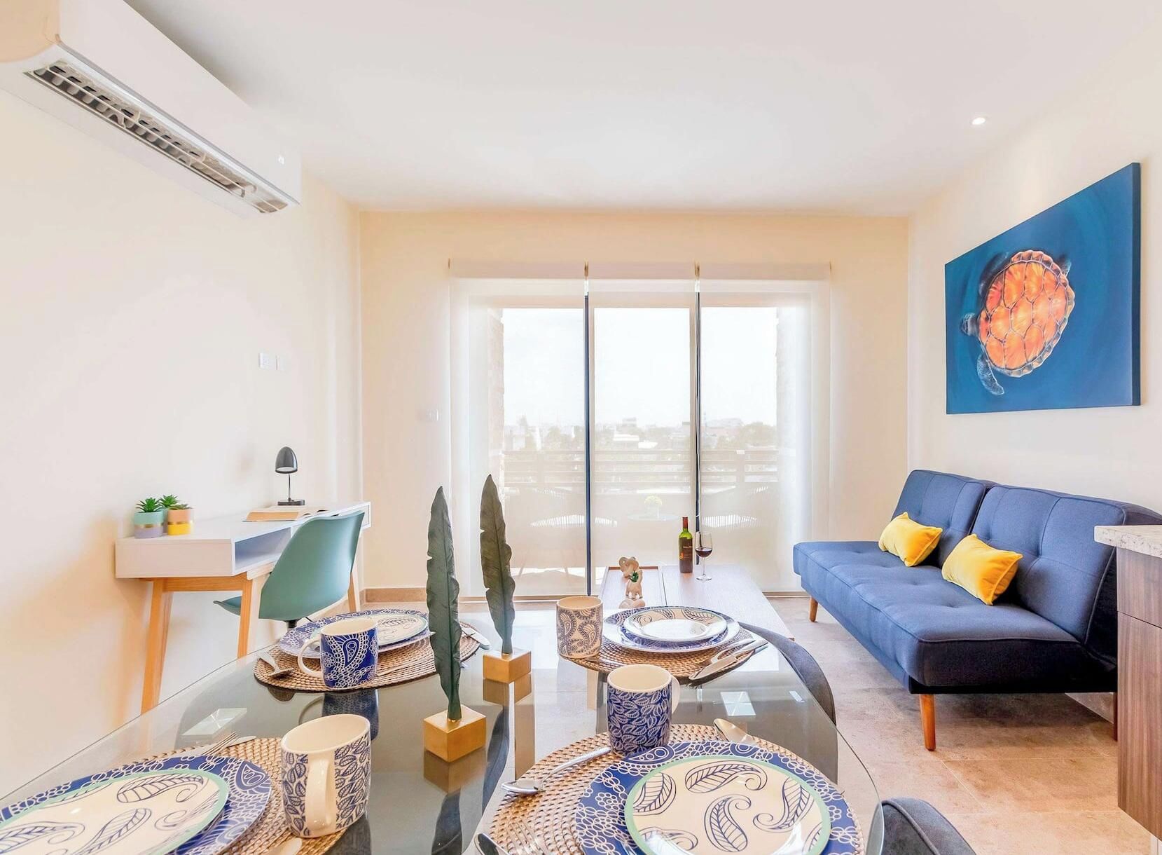 A modern living room with a glass dining table set for two, a blue sofa with yellow cushions, and a city view through large windows.