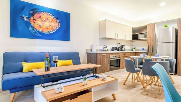 Una sala de estar moderna con un sofá azul, una mesa de centro de madera y una zona de cocina con armarios de madera y un refrigerador de acero inoxidable.