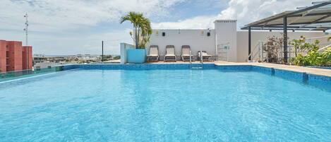 A clear blue swimming pool with a tiled edge, surrounded by lounge chairs and a palm tree.