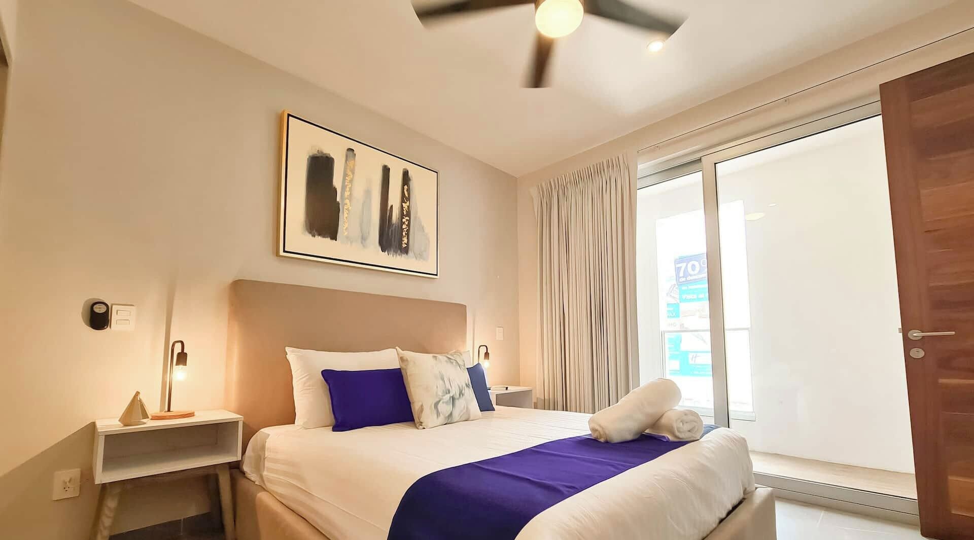 Ein Hotelzimmer mit Bett, Nachttisch, Deckenventilator, Fenster mit Vorhängen und einer Tür.