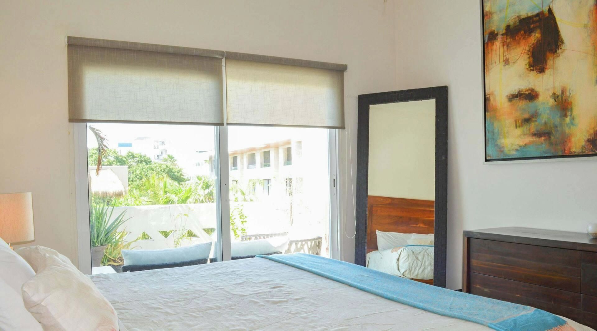 Une chambre d’hôtel avec un grand lit, une table de chevet, un miroir et une vue sur la verdure et les bâtiments environnants.