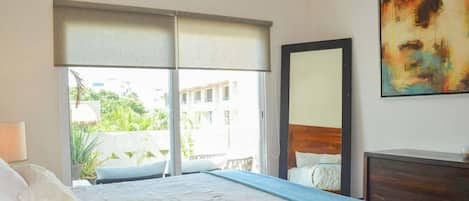 Une chambre d’hôtel avec un grand lit, une table de chevet, un miroir et une vue sur la verdure et les bâtiments environnants.