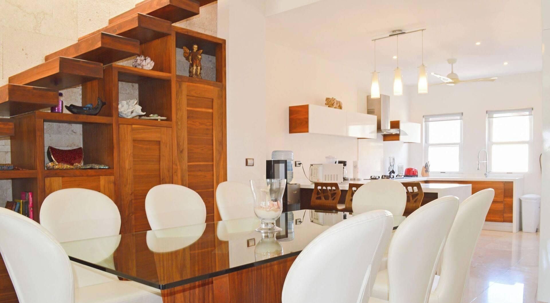 Una cocina moderna con armarios blancos, un área de comedor con mesa de vidrio y sillas blancas, y una escalera con barandillas de madera.