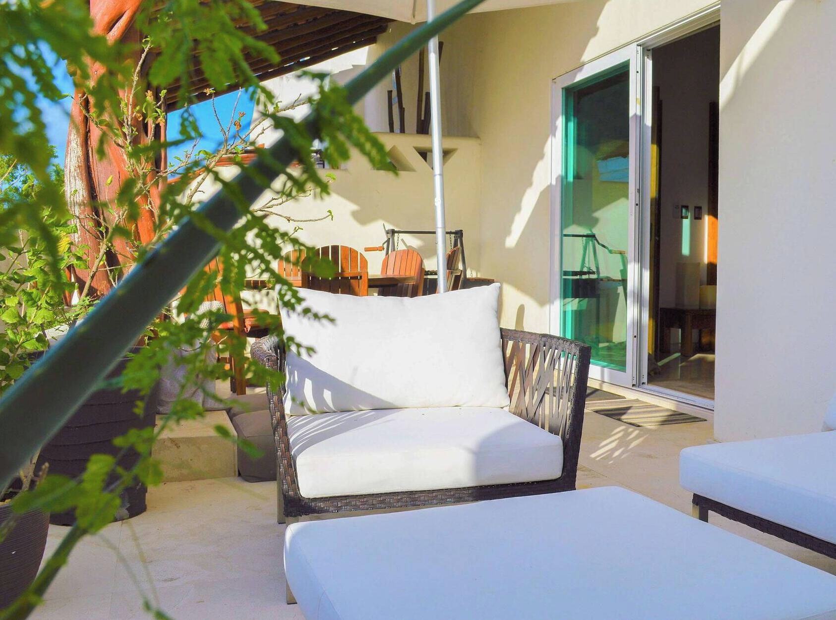 A patio with a wicker chair and a white cushion, a glass door leading inside, and a potted plant in the foreground.
