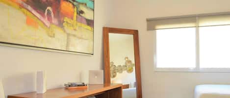 A hotel room with a bed, a desk with a mirror, a wall art piece, and a window with blinds.