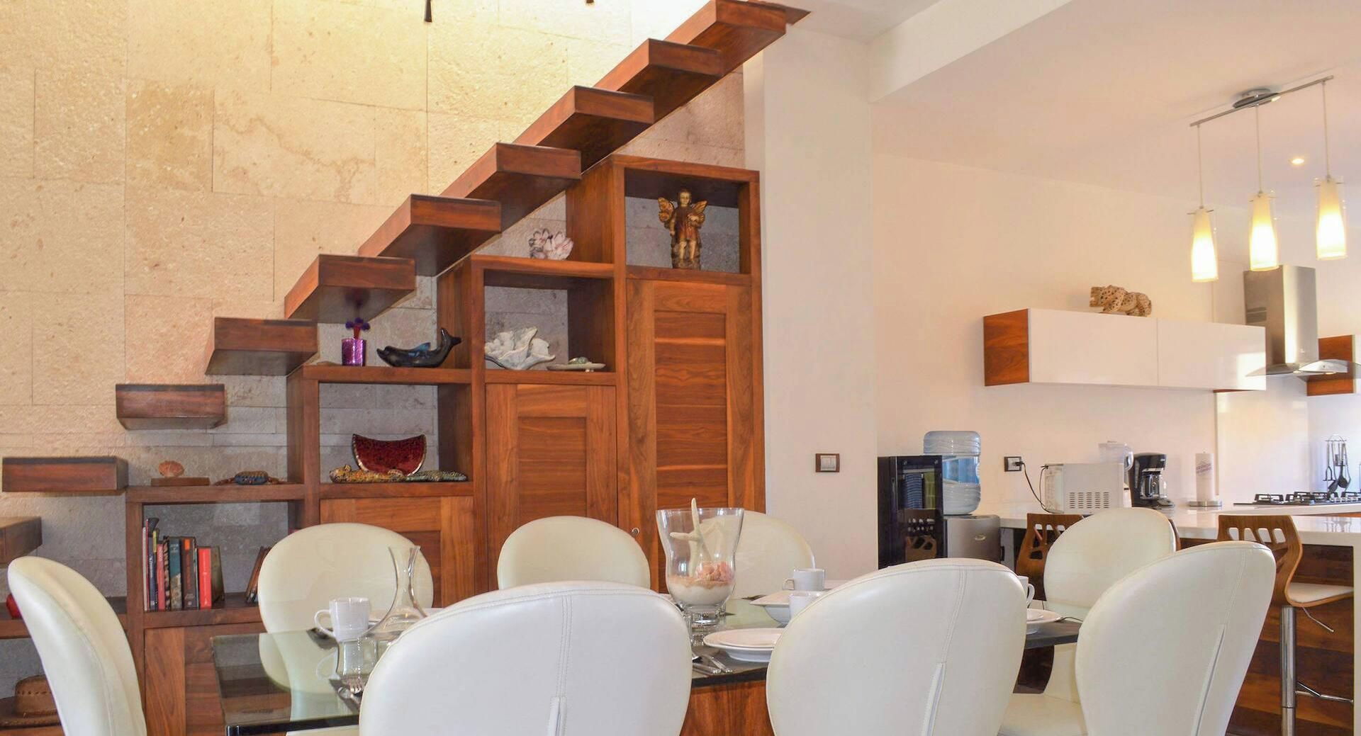 Un comedor con una escalera de madera, sillas blancas y una mesa dispuesta para cuatro.