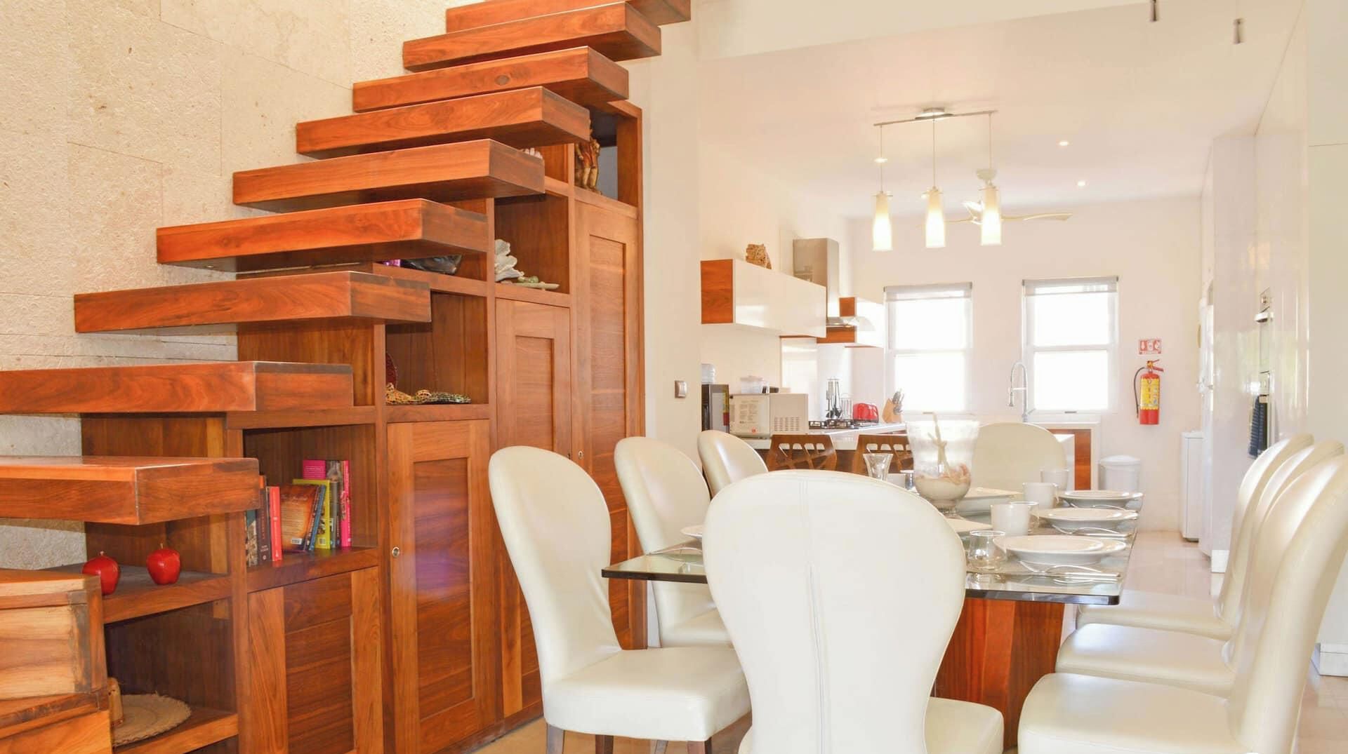A modern kitchen with a wooden staircase, white chairs, and pendant lights.