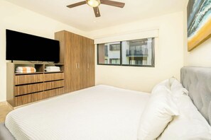 Een slaapkamer met een bed, een tv bevestigd aan een houten kast, een raam met uitzicht op gebouwen en een plafondventilator.