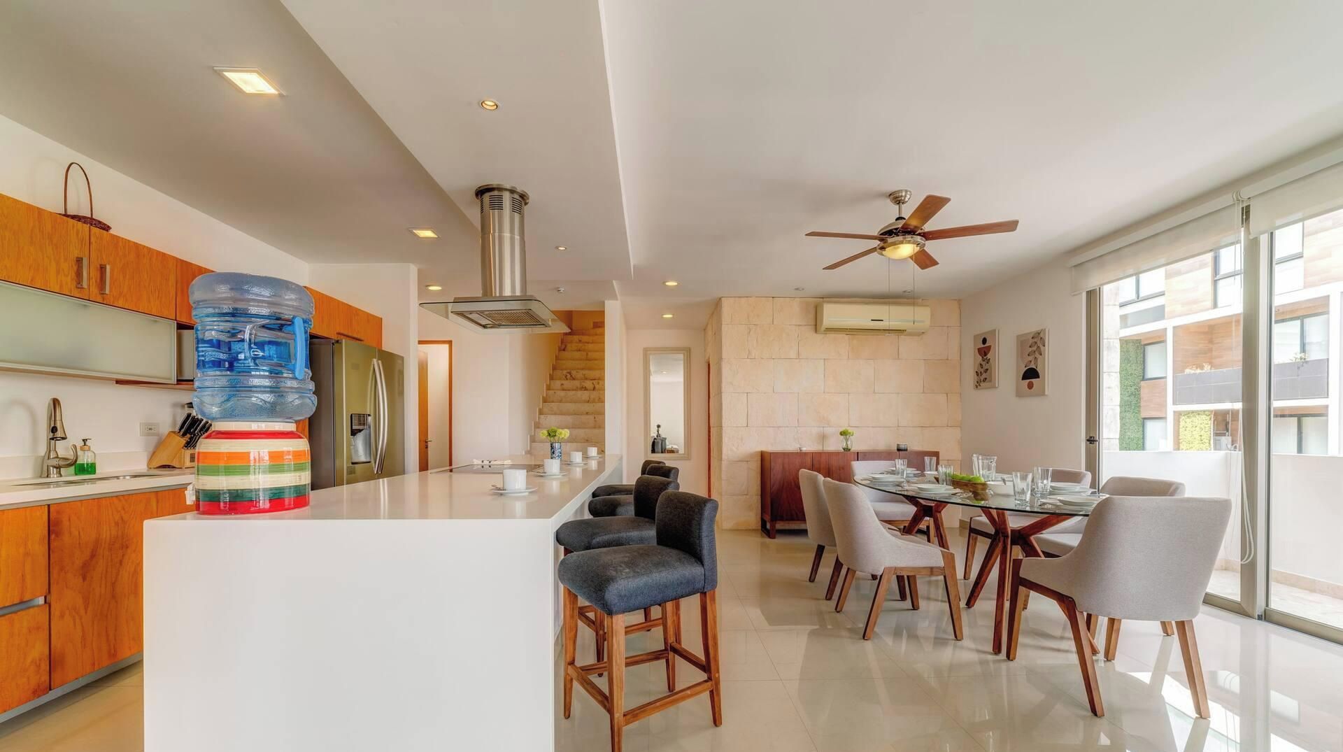 Une cuisine moderne dotée d’un îlot central, d’un coin repas avec une table en verre et d’un ventilateur de plafond.