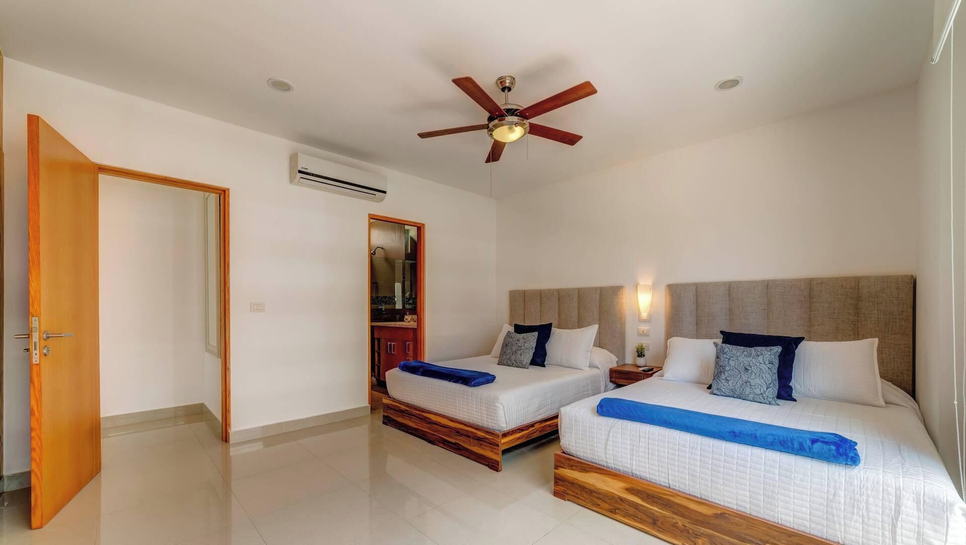 Een hotelkamer met twee bedden, een plafondventilator, een airconditioning en een houten deur.