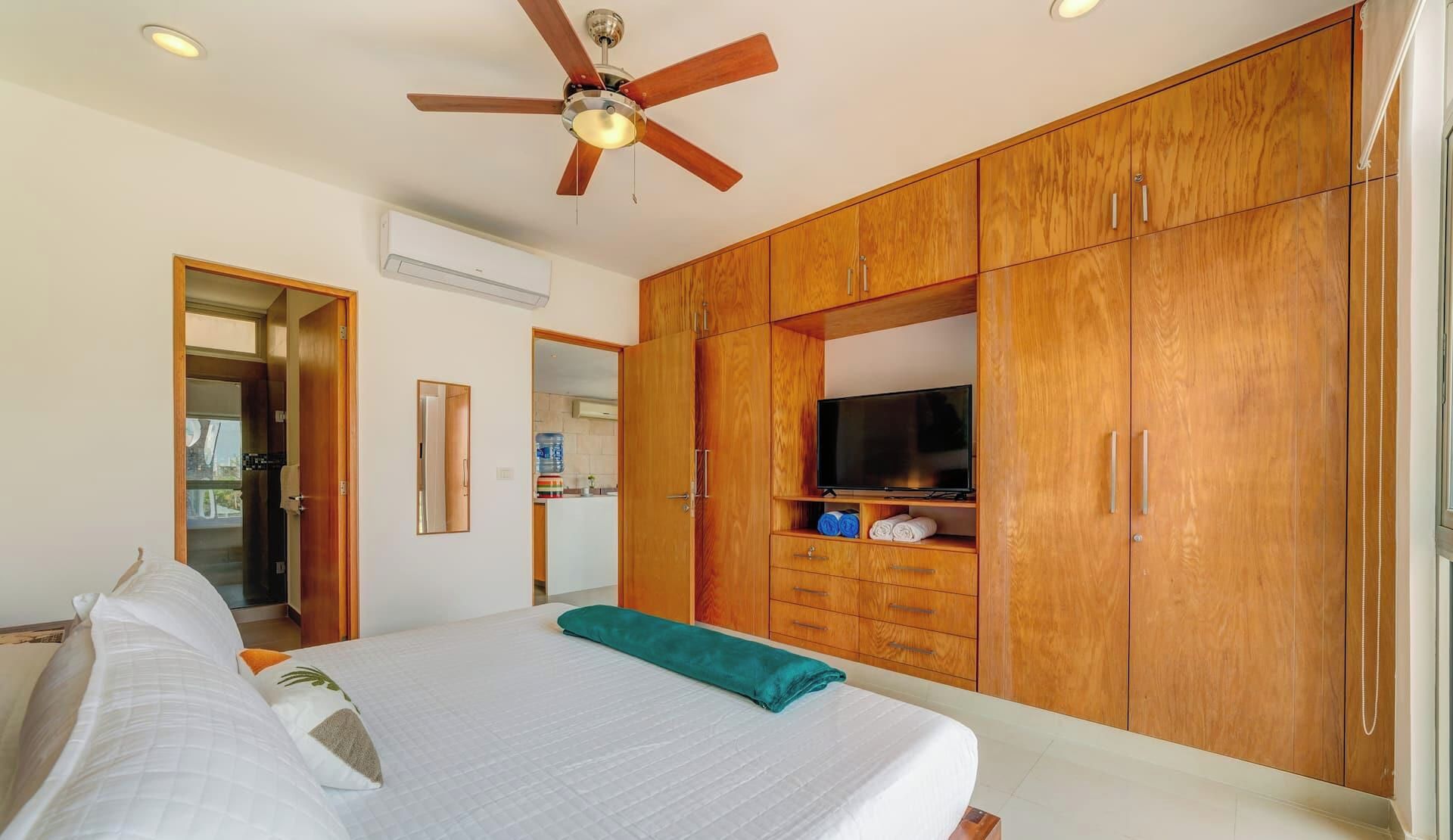 Quarto com cama, ventoinha no teto, guarda-roupa de madeira e televisão.