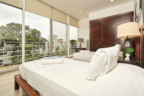 Ein Hotelzimmer mit einem großen Bett, einem Holzschrank, einer Nachttischlampe und Blick auf die Stadt durch große Fenster.