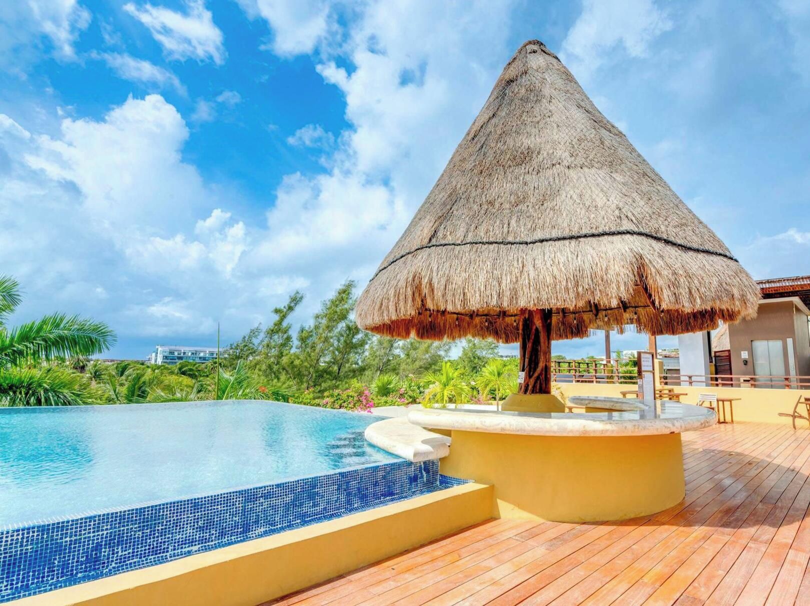 A swimming pool with a thatched umbrella, wooden deck, and a view of the ocean.