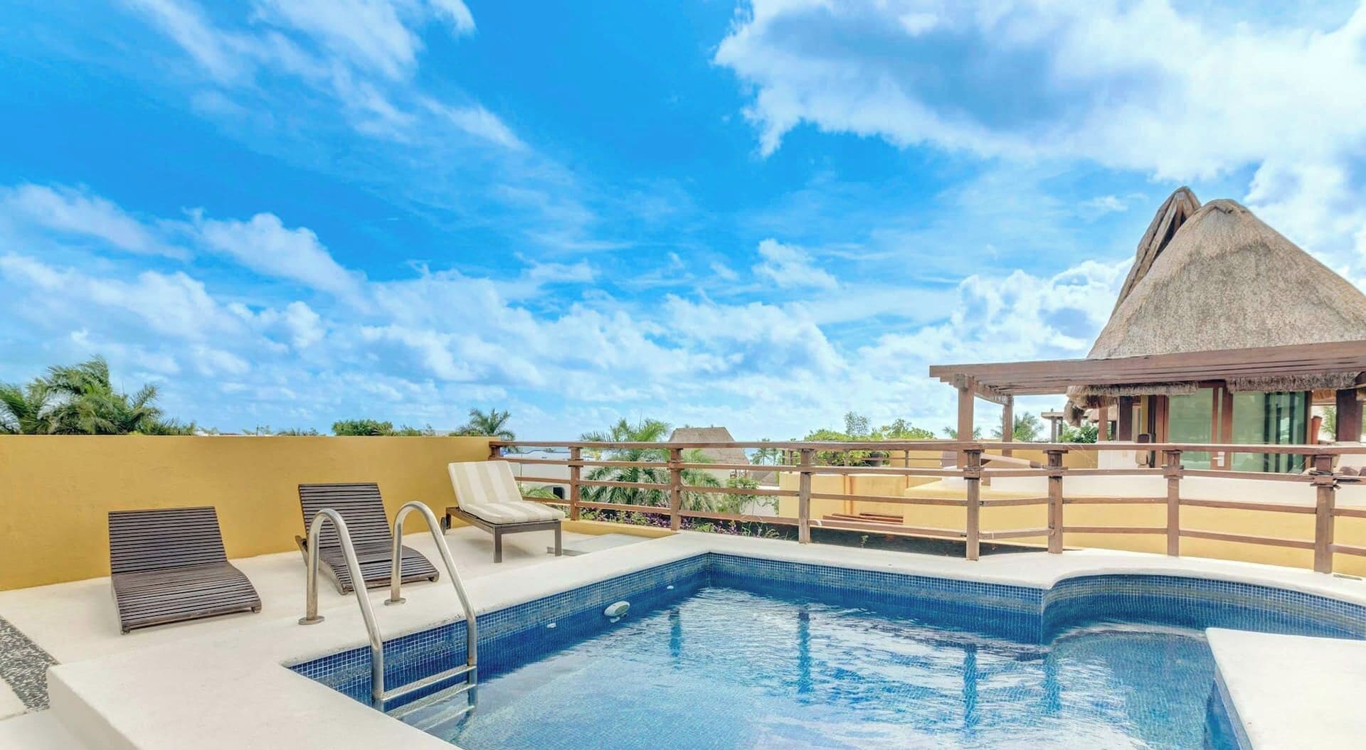 Una piscina en la azotea con vistas despejadas del cielo, un edificio con techo de paja y un área de descanso con sillones.