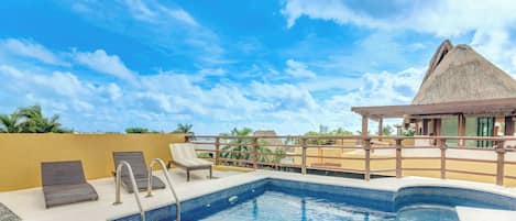 Una piscina en la azotea con vistas despejadas del cielo, un edificio con techo de paja y un área de descanso con sillones.