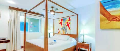 Una cama con dosel, un ventilador de techo, una ventana con cortinas, una mesita de noche con lámpara y un cuadro en la pared.