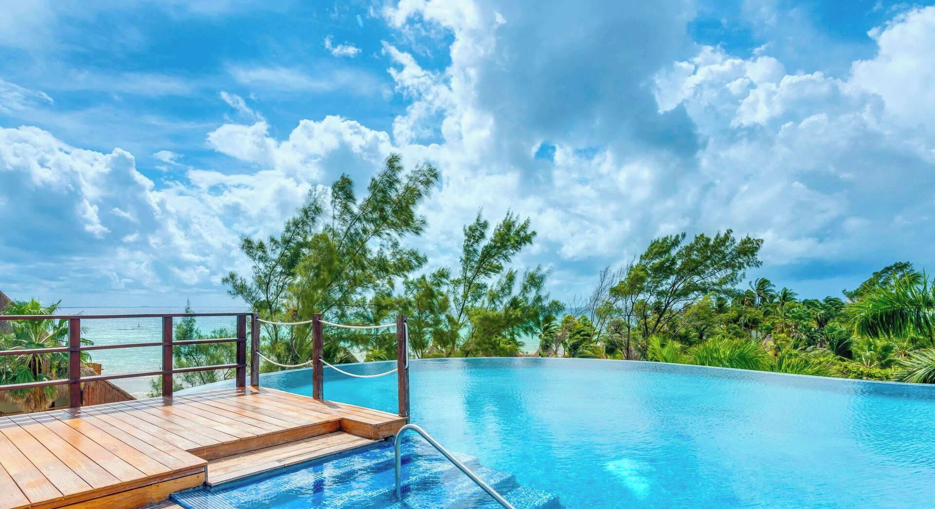 A clear blue sky with clouds, a wooden deck, and a swimming pool surrounded by lush greenery.