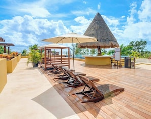Een houten terras met lounge fauteuils, een rieten zonnescherm en een zwembadgedeelte met uitzicht op de oceaan.