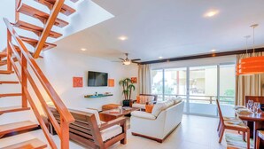 A modern living room with a wooden staircase, a flat-screen TV, a sofa, and a dining area with a table set for two. - Penthouse w/ private terrace & rooftop pool (Playa del Carmen)