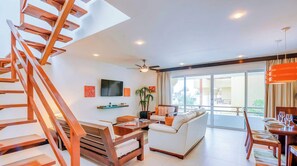 A modern living room with a wooden staircase, a flat-screen TV, a sofa, and a dining area with a table set for two. - Penthouse w/ private terrace & rooftop pool (Playa del Carmen)