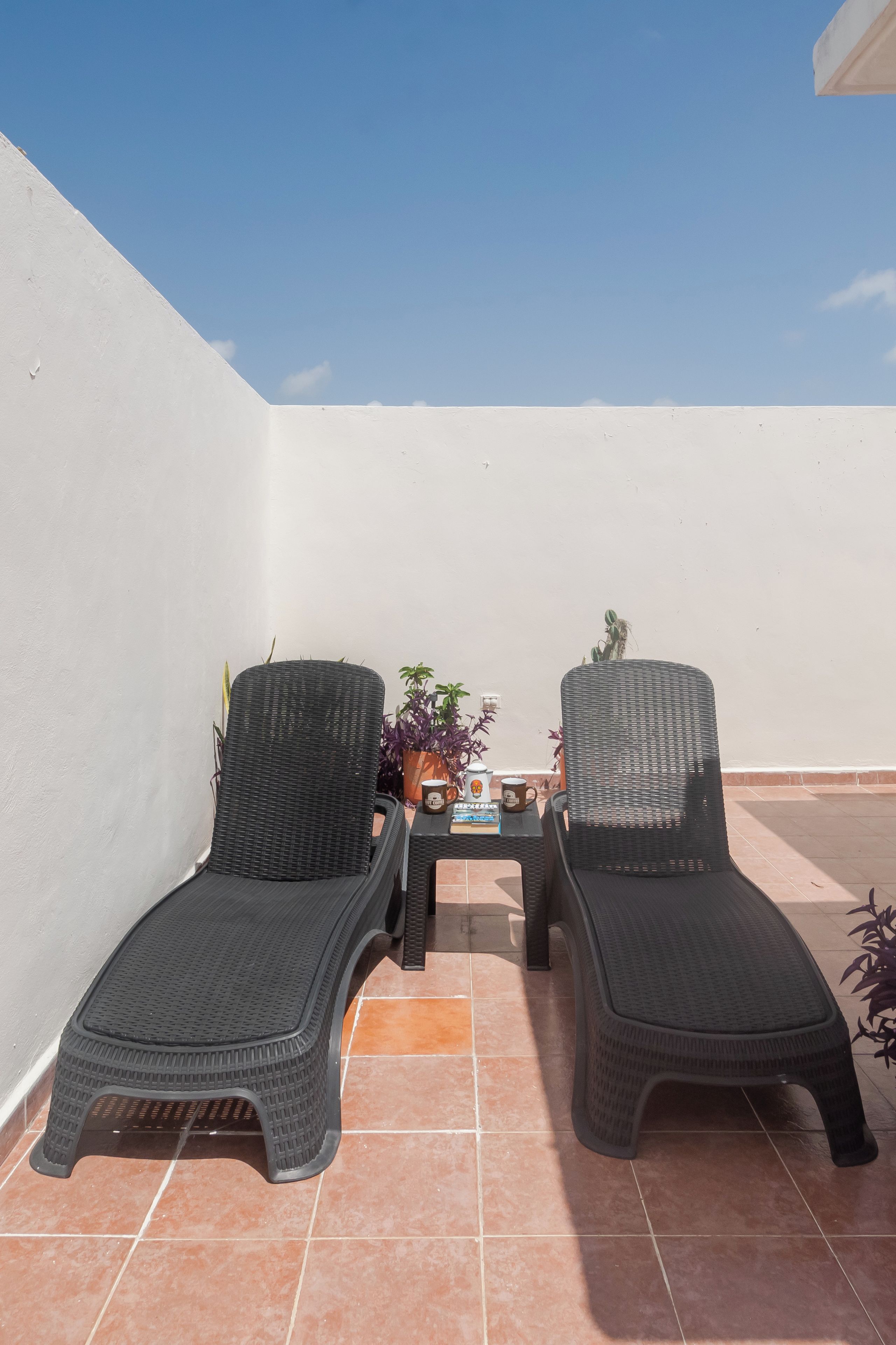 Two wicker lounge chairs on a tiled patio with a white wall and a clear blue sky in the background.