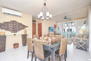A dining area with a glass table, woven chairs, a chandelier, and a wall-mounted air conditioner. - 2BR condo - pool, BBQ & beach steps away (Playa del Carmen)