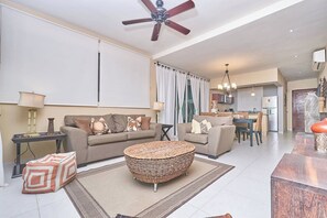 A modern living room with a sofa, a round wicker ottoman, a dining table with chairs, a ceiling fan, and a kitchen area in the background. - 2BR condo - pool, BBQ & beach steps away (Playa del Carmen)
