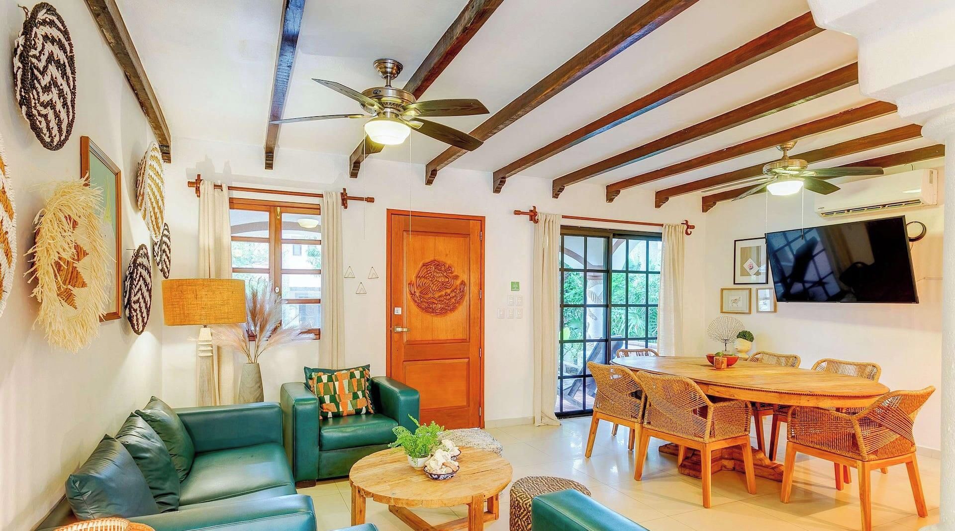 A spacious living area with a green sofa, a wooden dining table with wicker chairs, a ceiling fan, and a TV mounted on the wall.