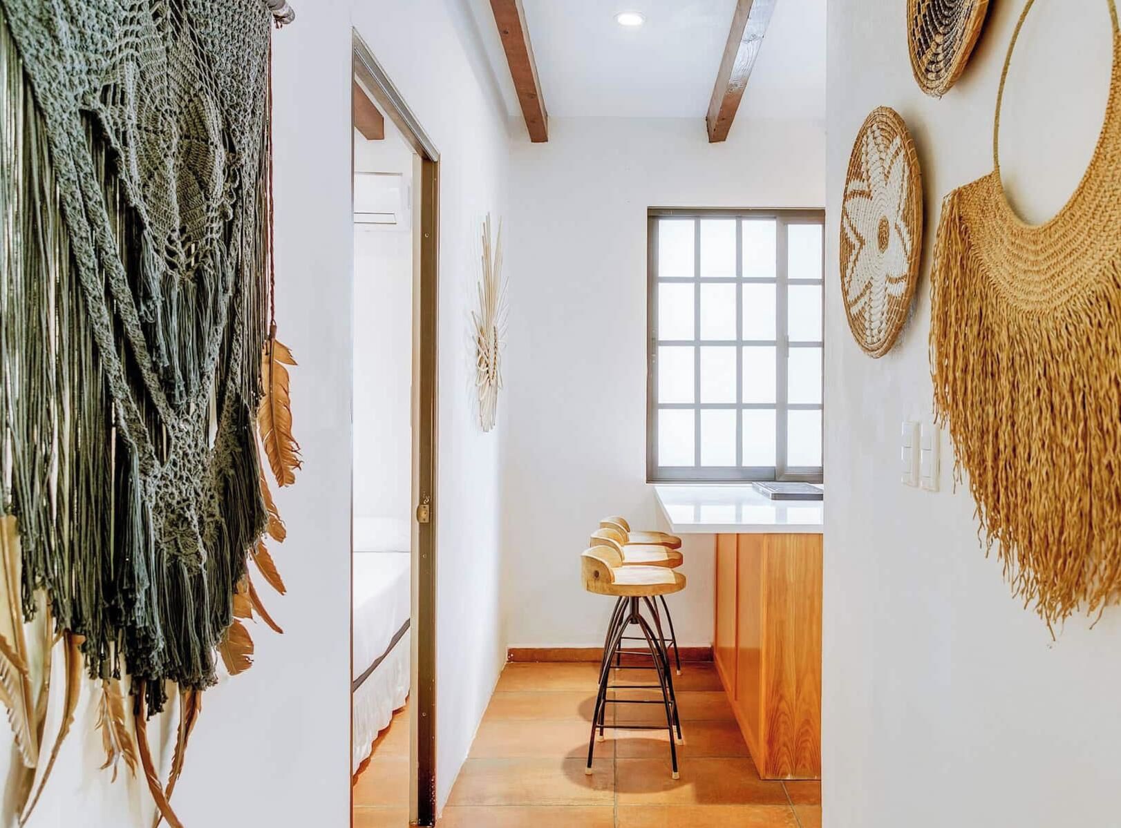 Una habitación con techo de madera, una ventana con persianas, un taburete de madera y un muro decorado con detalles tejidos.